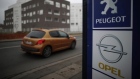 A Peugeot car drives past the logos of French car maker Peugeot and German car maker Opel